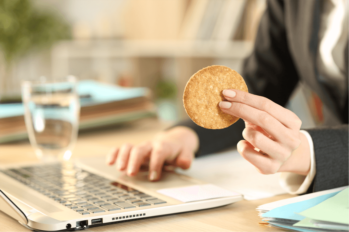 A person typing on a laptop while holding a cookie.